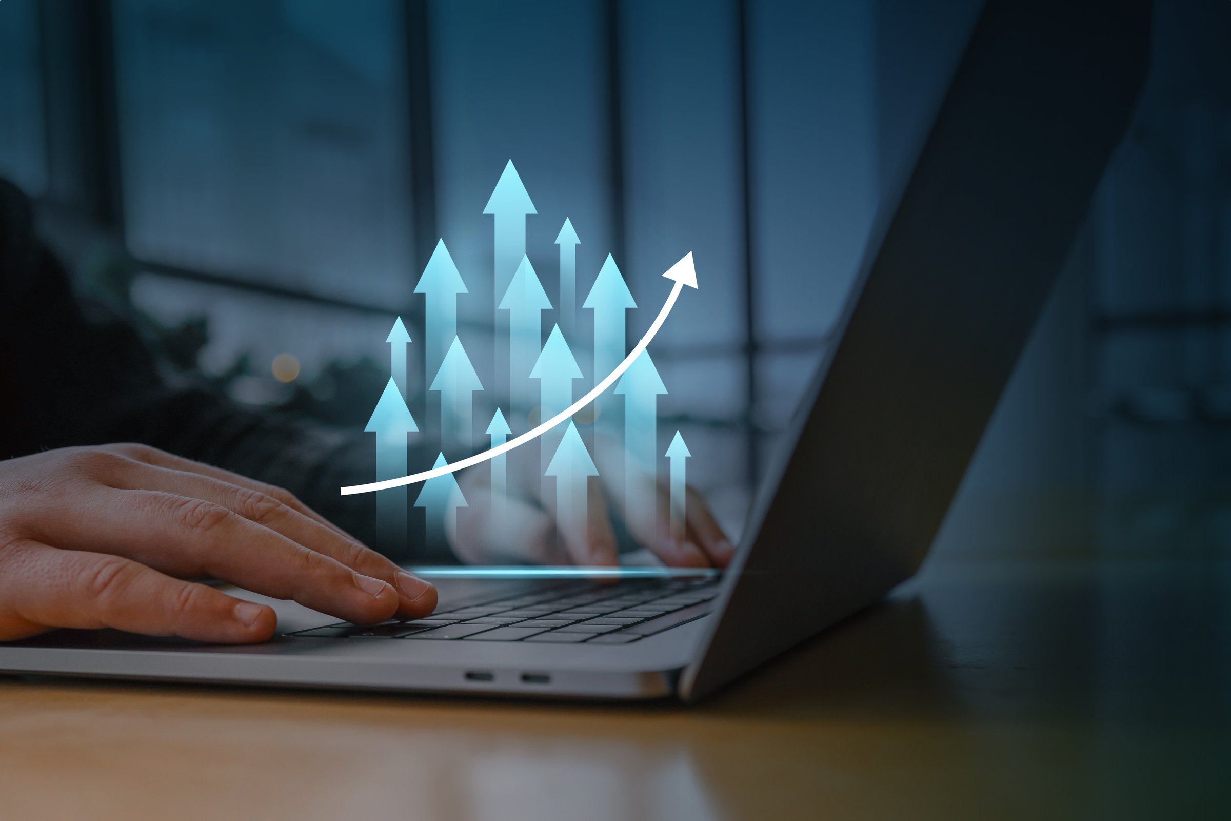 A person working on a laptop with a digital overlay of glowing upward arrows and a rising graph. Concept of business growth, success, financial progress, and digital marketing strategy.